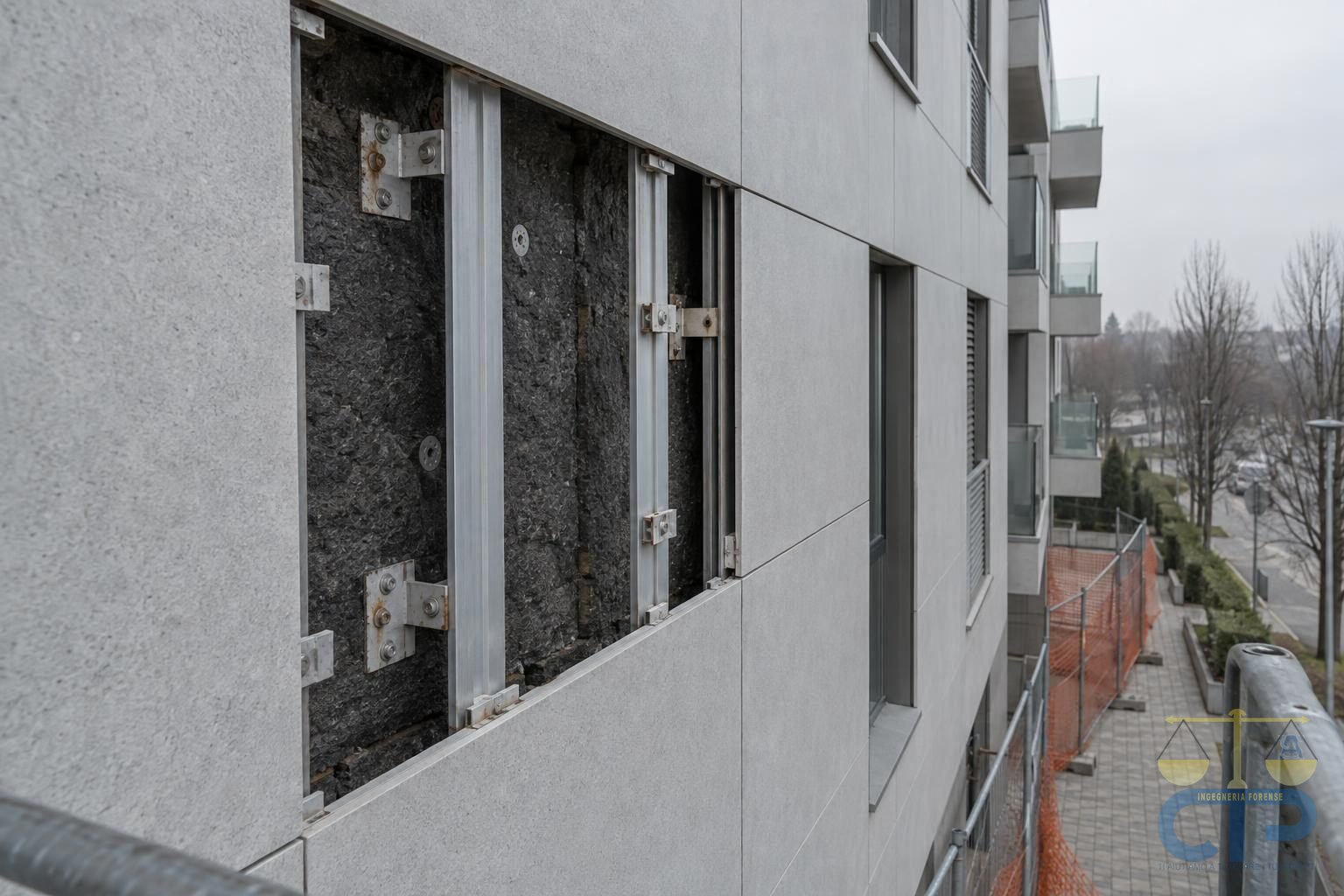 Facciata ventilata di un edificio residenziale recente con una lastra di rivestimento mancante: si vedono i montanti in alluminio, le staffe di ancoraggio e l'isolante scuro nell'intercapedine; sotto, rete di protezione arancione sul marciapiede